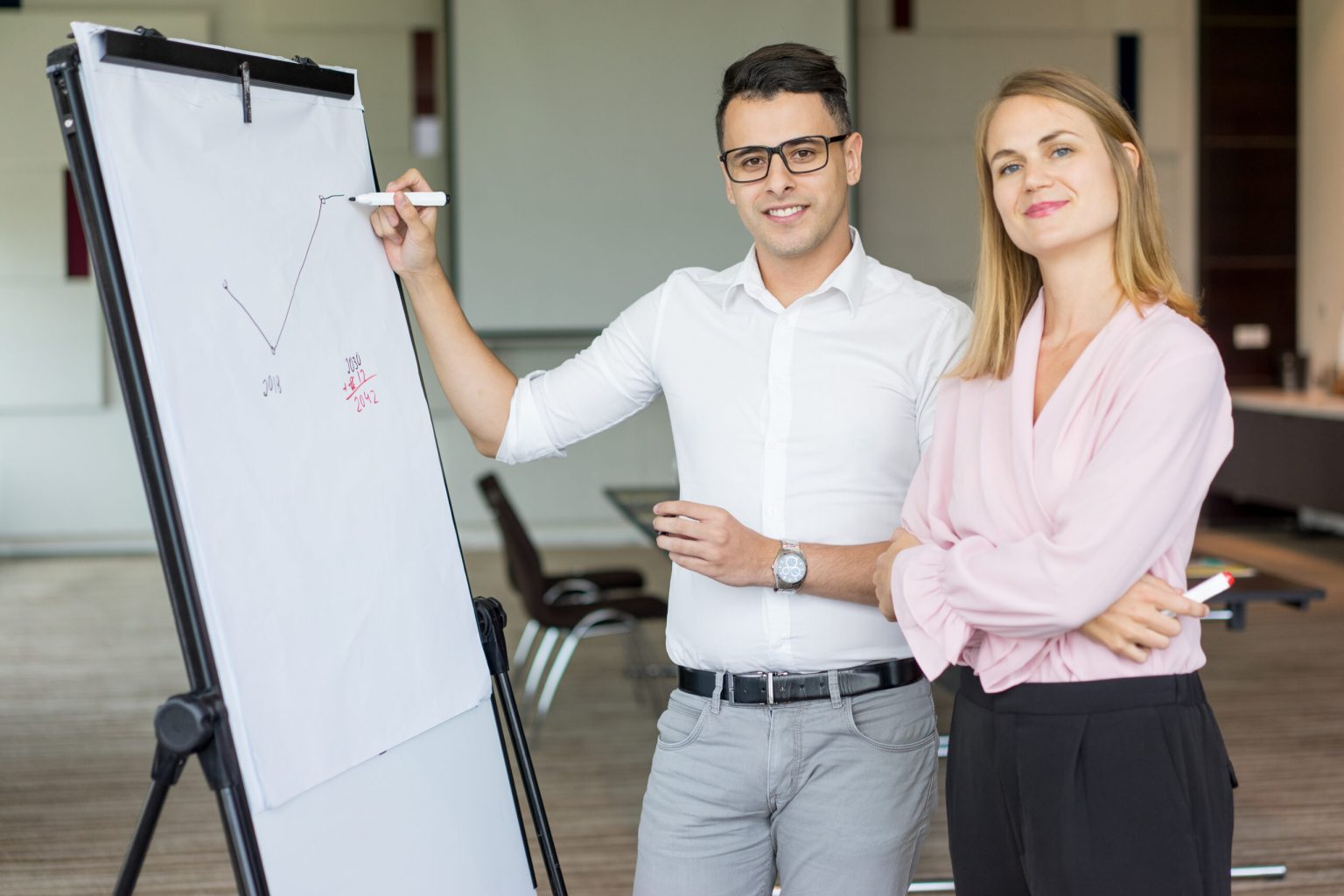 Portrait of smiling business colleagues drawing plan on flipchart. Young Caucasian businessman wearing glasses and businesswoman smiling at camera in boardroom. Teamwork and briefing concept